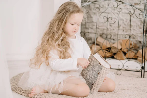 Linda menina bonito segurando um livro e sentado perto de lareira branca dentro de casa sozinho — Fotografia de Stock