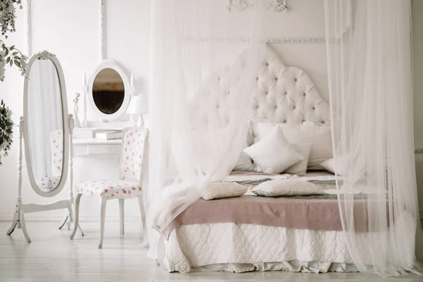 Biała sypialnia wnętrze z nikt. duże wygodne łóżko z białym baldachimem i owalne lustro toaletka z toaletką obok niego — Zdjęcie stockowe