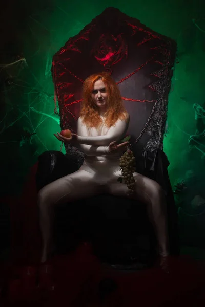 Beautiful Young plus size red hair woman as seductive temptress. halloween portrait of evil curvy woman in skin color outfit on black gothic throne in the dark in green light. — Stock Photo, Image