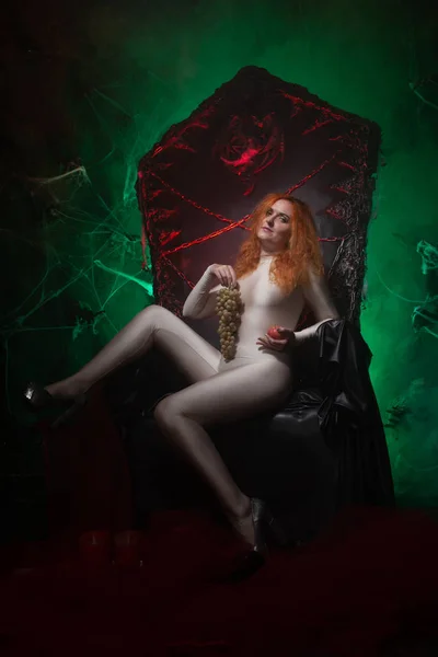 Beautiful Young plus size red hair woman as seductive temptress. halloween portrait of evil curvy woman in skin color outfit on black gothic throne in the dark in green light. — Stock Photo, Image