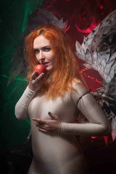 Mysterious plus size red hair girl in skincolor beige outfit with black and white big demon wings on gothic cemetery. Witch or dark angel style. Woman tempts by apple in the middle of the night — Stock Photo, Image