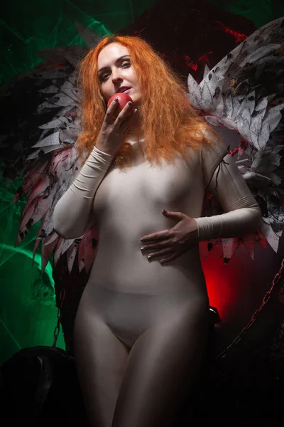 Mysterious plus size red hair girl in skincolor beige outfit with black and white big demon wings on gothic cemetery. Witch or dark angel style. Woman tempts by apple in the middle of the night — Stock Photo, Image