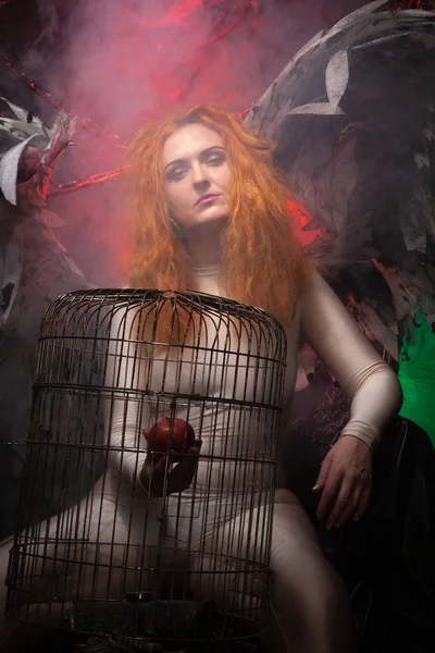 Zła kusząca kobieta z dużymi skrzydłami demonów trzyma jabłko w dużej klatce i kusi grzechem. Halloween zdjęcie plus rozmiar dziewczyna z czerwonymi włosami na ogromnym tronie gotyckim. — Zdjęcie stockowe