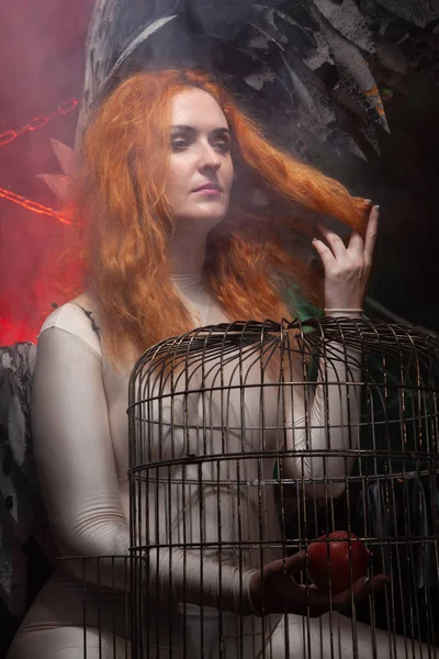 An evil tempting woman with large demon wings holds an Apple in a large cage and beckons to sin. Halloween photo plus size girl with red hair on a huge Gothic throne. — Stock Photo, Image