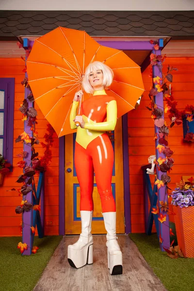 Mujer rubia bonita caliente en traje de goma de látex colorido brillante listo para la fiesta de otoño de Halloween y posando con paraguas en forma de corazón cerca de su casa naranja —  Fotos de Stock