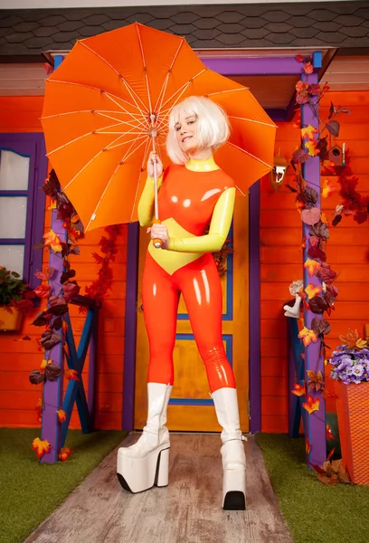 Hot pretty blonde woman in bright colorful latex rubber catsuit ready for halloween autumn party and posing with heart shape umbrella near her orange house — Stock Photo, Image