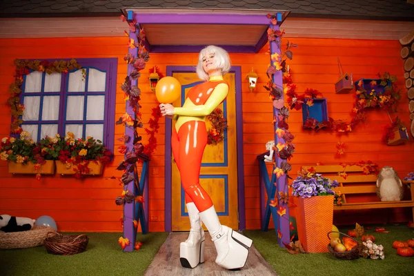 Hete mooie blonde vrouw in heldere kleurrijke latex rubber Catsuit klaar voor Halloween herfst feest en poseren met lucht ballon in de buurt van haar oranje huis — Stockfoto