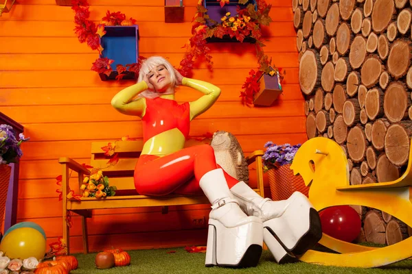 Mujer rubia bonita caliente en traje de goma de látex colorido brillante listo para la fiesta de otoño de Halloween y posando cerca de su casa naranja —  Fotos de Stock