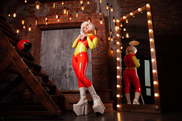 модная женщина в тесном пространстве яркий комбинезон с белой платформой сапоги позирует рядом с огромным зеркалом пола с лампочками дома в одиночку. тело в полный рост
.