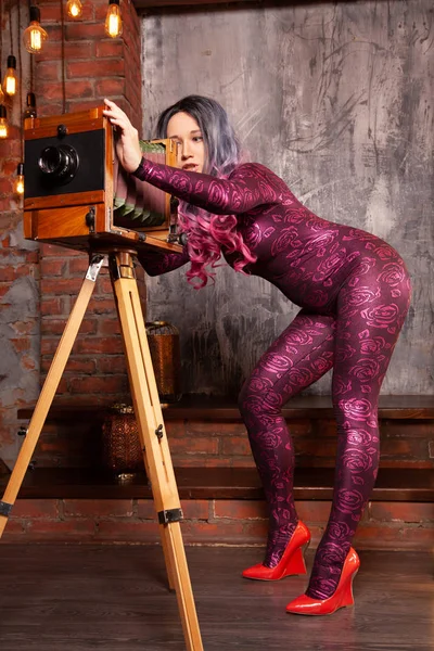 Charming luxury fashionable woman in a tight jump suit with a retro large camera posing and taking pictures among the bright light bulbs — Stock Photo, Image