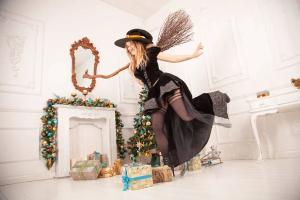 La bruja malvada en vestido negro y sombrero de miedo de Halloween vino para Navidad y trata de estropear una fiesta, balanceando la vieja escoba entre una hermosa habitación con un árbol de Navidad con regalos —  Fotos de Stock