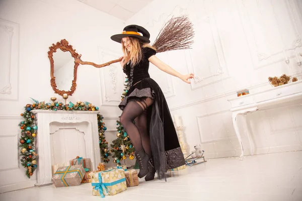 Zła wiedźma w czarnej sukience i straszny kapelusz z Halloween przyszedł na Boże Narodzenie i próbuje zepsuć wakacje, huśtawka starej miotły wśród pięknego pokoju z choinką z prezentami — Zdjęcie stockowe