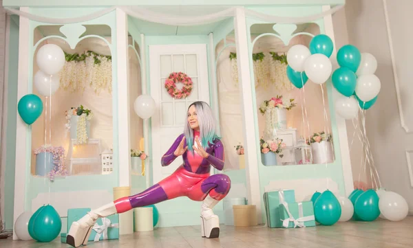 Vrij schattig volwassen meisje in paars latex rubber kostuum poseren in de buurt van cadeauwinkel met cadeautjes en luchtballonnen. fetisj vrouw in strakke outfit poseren alleen. volledige lengte portret. — Stockfoto