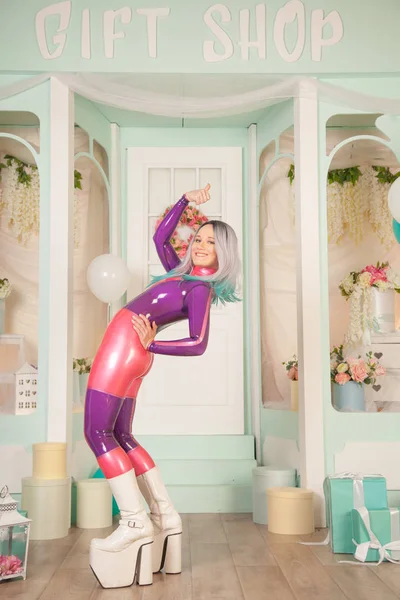 Vrij schattig volwassen meisje in paars latex rubber kostuum poseren in de buurt van cadeauwinkel met cadeautjes en luchtballonnen. fetisj vrouw in strakke outfit poseren alleen. volledige lengte portret. — Stockfoto