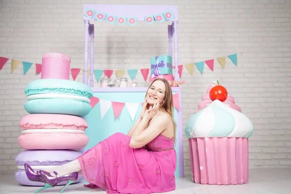 Linda chica adulta feliz posando en vestido de moda entre enormes dulces y pasteles fabulosos —  Fotos de Stock