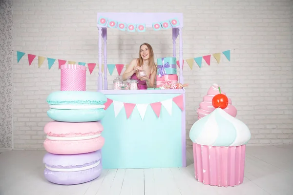 Linda chica adulta feliz posando en vestido de moda entre enormes dulces y pasteles fabulosos —  Fotos de Stock