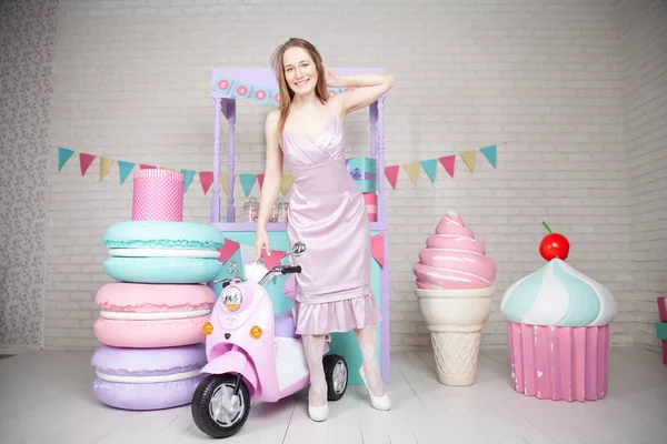 Linda chica adulta feliz posando en vestido de moda entre enormes dulces y pasteles fabulosos —  Fotos de Stock