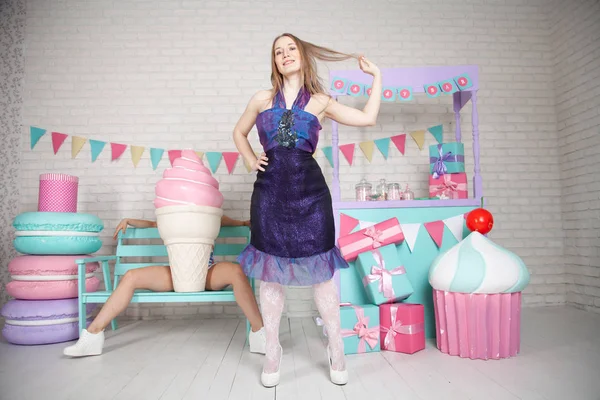 Linda chica adulta feliz posando en vestido de moda entre enormes dulces y pasteles fabulosos — Foto de Stock