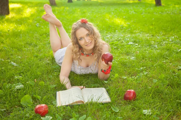 Blank gelukkig student meisje in schattig wit jurk rusten op de groene zomer gras met boek en rode appels — Stockfoto