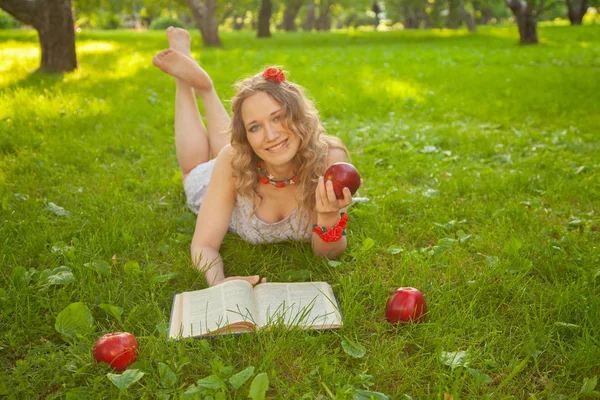 Caucasiano menina estudante feliz em bonito vestido branco resto na grama de verão verde com livro e maçãs vermelhas — Fotografia de Stock