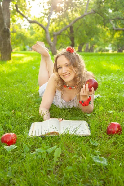 Caucasiano menina estudante feliz em bonito vestido branco resto na grama de verão verde com livro e maçãs vermelhas — Fotografia de Stock