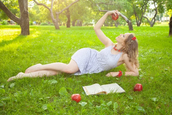 Caucásico feliz estudiante chica en lindo vestido blanco resto en la hierba verde de verano con libro y manzanas rojas — Foto de Stock