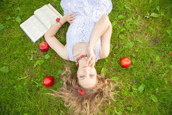 Caucasiano menina estudante feliz em bonito vestido branco resto na grama de verão verde com livro e maçãs vermelhas — Fotografia de Stock