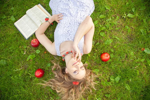 Caucasiano menina estudante feliz em bonito vestido branco resto na grama de verão verde com livro e maçãs vermelhas — Fotografia de Stock