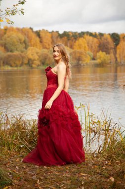 Güz bahçesinde gölün kenarında poz veren uzun gece kıyafetli genç bir kadın.