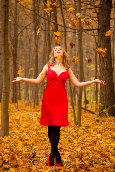 Ein Mädchen im roten Kleid wirft allein im herbstlichen Park mit Blättern — Stockfoto