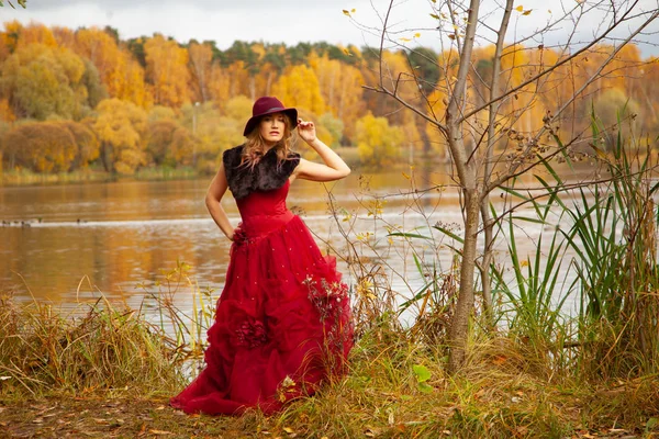 Młoda kobieta w stylowej czapce i długiej czerwonej sukni wieczorowej z futra pozowanie w pobliżu rzeki i chodzenie w jesiennym tle ogrodu sam — Zdjęcie stockowe