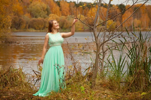 Junge Frau in mintblauem langen Kleid steht allein am Wasser im herbstlichen Gartenhintergrund — Stockfoto