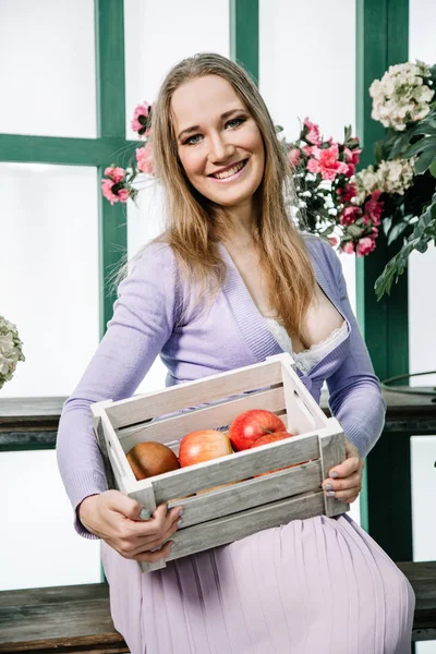 Beautiful girl in a purple dress with a box of apples in her hands in the greenhouse poses among the plants and enjoys the clean air and harmony of communication with nature — 스톡 사진