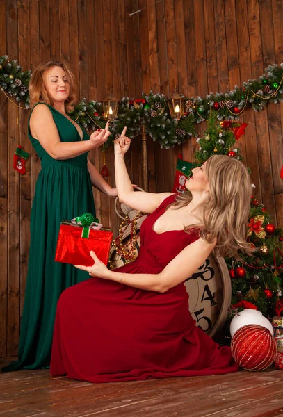 Due donne eleganti in abito da sera lungo rosso e verde in posa vicino all'albero di Natale al chiuso — Foto Stock