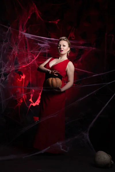 Woman in evening classic dress posing with pumpkin on black Halloween background with spider web — Stock Photo, Image