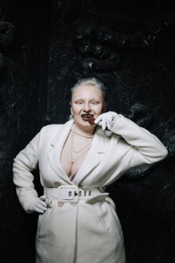 Genç ucube artı boyut albino kadın açgözlülükle yiyor ve siyah arka planda çikolatayı seviyor. kıvrımlı kısa saçlı beyaz elbiseli kız..