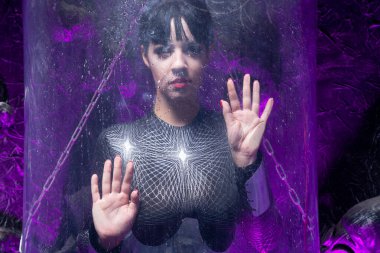 Cam uzay kapsülünün yanında poz veren fütüristik bir gencin fotoğrafı. Siyah arka planda mor neon ışığı ve kablolar var.