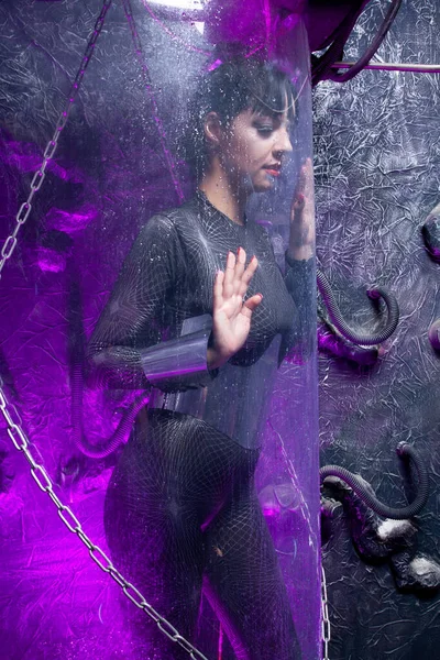 Fotografía de una joven futurista posando cerca de una cápsula espacial de vidrio con cables y luz de neón púrpura sobre fondo negro —  Fotos de Stock