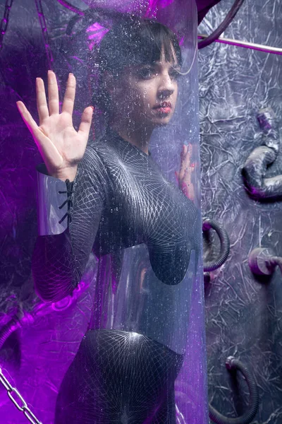 Fotografía de una joven futurista posando cerca de una cápsula espacial de vidrio con cables y luz de neón púrpura sobre fondo negro —  Fotos de Stock