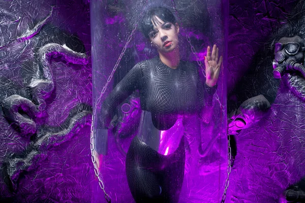 Fotografía de una joven futurista posando cerca de una cápsula espacial de vidrio con cables y luz de neón púrpura sobre fondo negro — Foto de Stock
