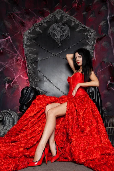スタジオで大きな黒い玉座の近くに赤い長いドレスの美しい吸血鬼の女性 — ストック写真