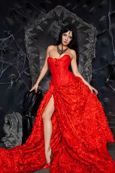 붉은 긴 옷을 입은 아름다운 뱀파이어 여인 이 스튜디오의 큰 검은 왕좌 근처에 있다 — 스톡 사진