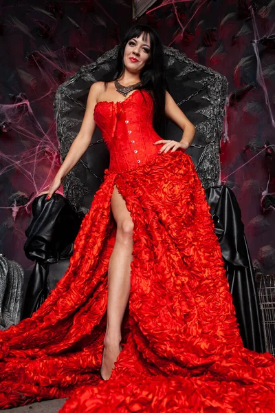 スタジオで大きな黒い玉座の近くに赤い長いドレスの美しい吸血鬼の女性 — ストック写真