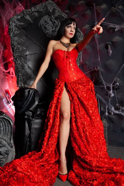 Beautiful vampire woman in red long dress near big black throne in the studio — Stock Photo, Image