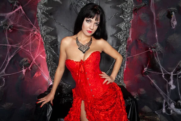 Красивая женщина вампир в красном длинном платье рядом с большим черным троном в студии — стоковое фото