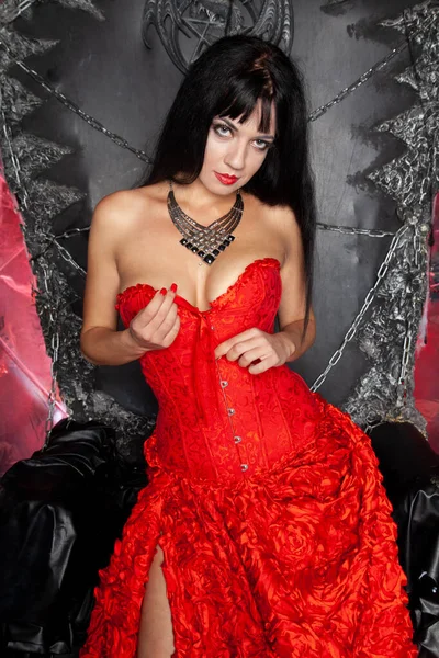 Bella donna vampiro in abito rosso lungo vicino grande trono nero in studio — Foto Stock
