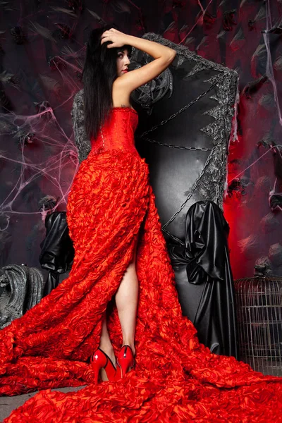 在演播室里穿着红色长裙的美丽吸血鬼女人接近黑色的大王座 — 图库照片