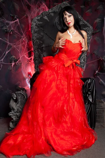 在演播室里穿着红色长裙的美丽吸血鬼女人接近黑色的大王座 — 图库照片