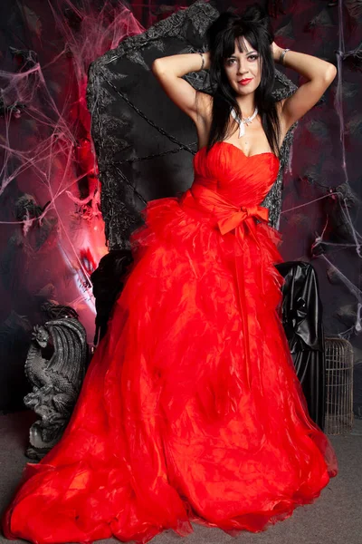 Beautiful vampire woman in red long dress near big black throne in the studio — Stock Photo, Image