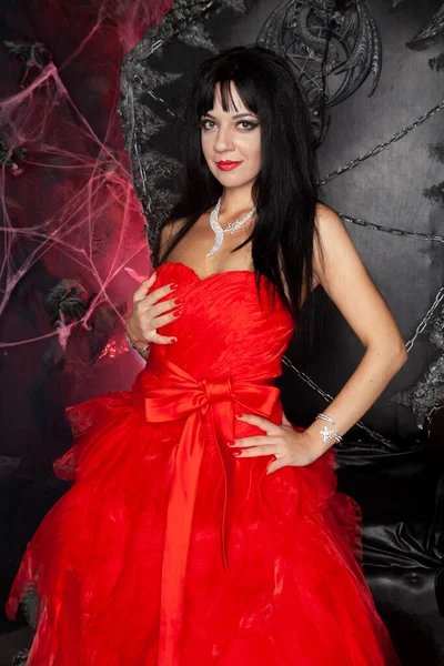 Bela mulher vampira em vestido longo vermelho perto de grande trono preto no estúdio — Fotografia de Stock